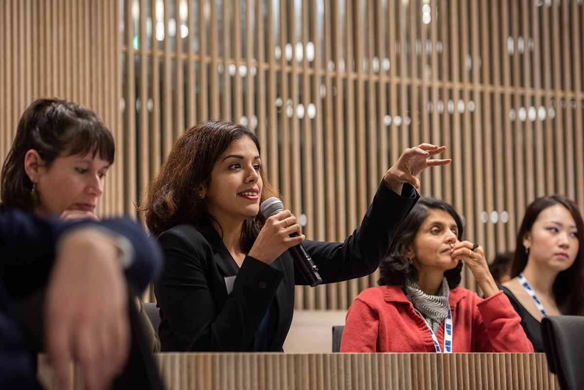 During a conference, a woman asks a question with a microphone in her right hand and raises her left hand