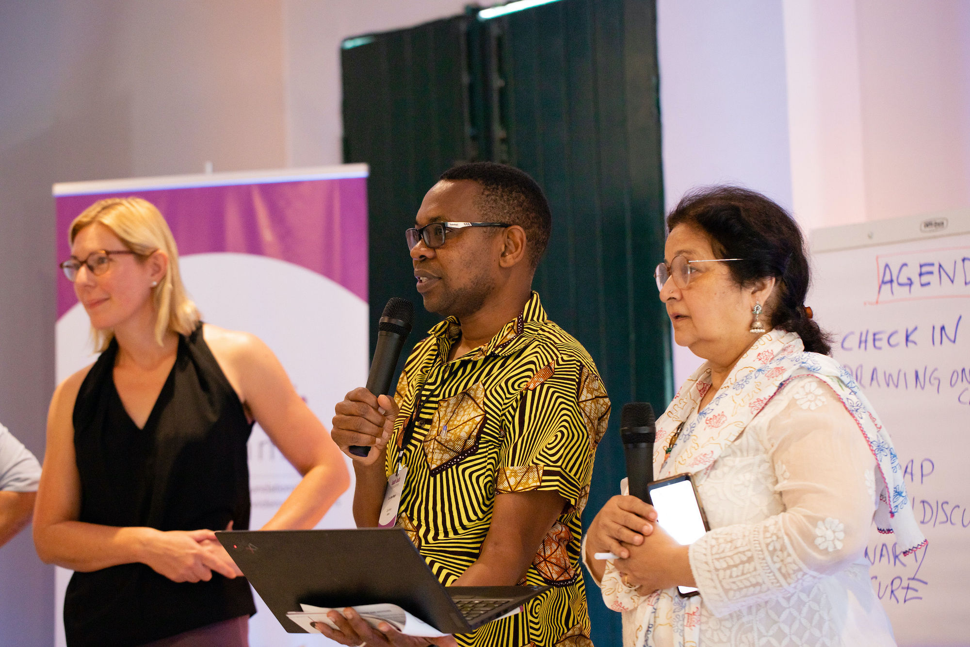 Man presents with a laptop and microphone. Women stand either side.