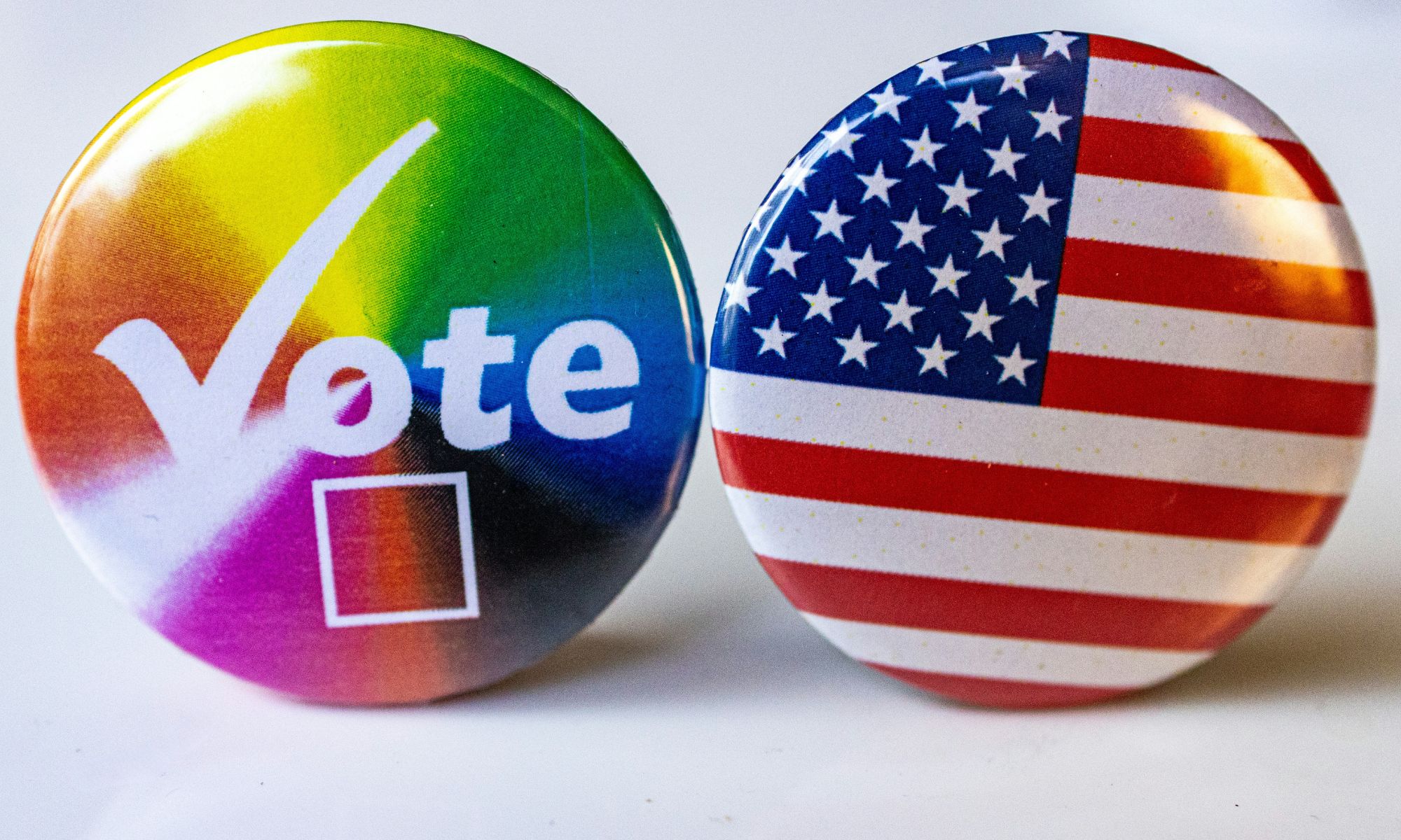 Two round badges side by side. The left badge has a rainbow background with the word "Vote" in white letters and a check box below it, symbolising LGBTQ+ pride and voting. The right badge features the design of the United States flag.