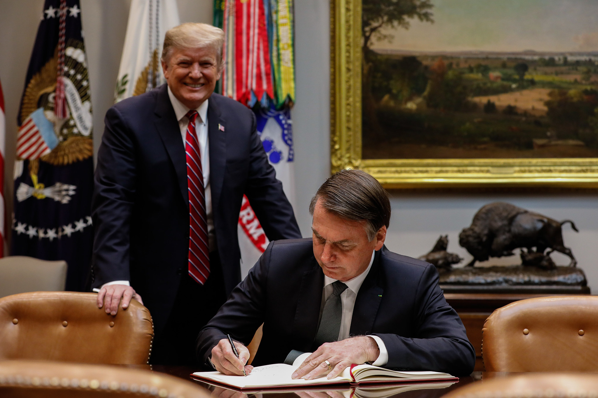 President of Brazil sits at a desk signing a document. President Trump is stood behind him smiling.