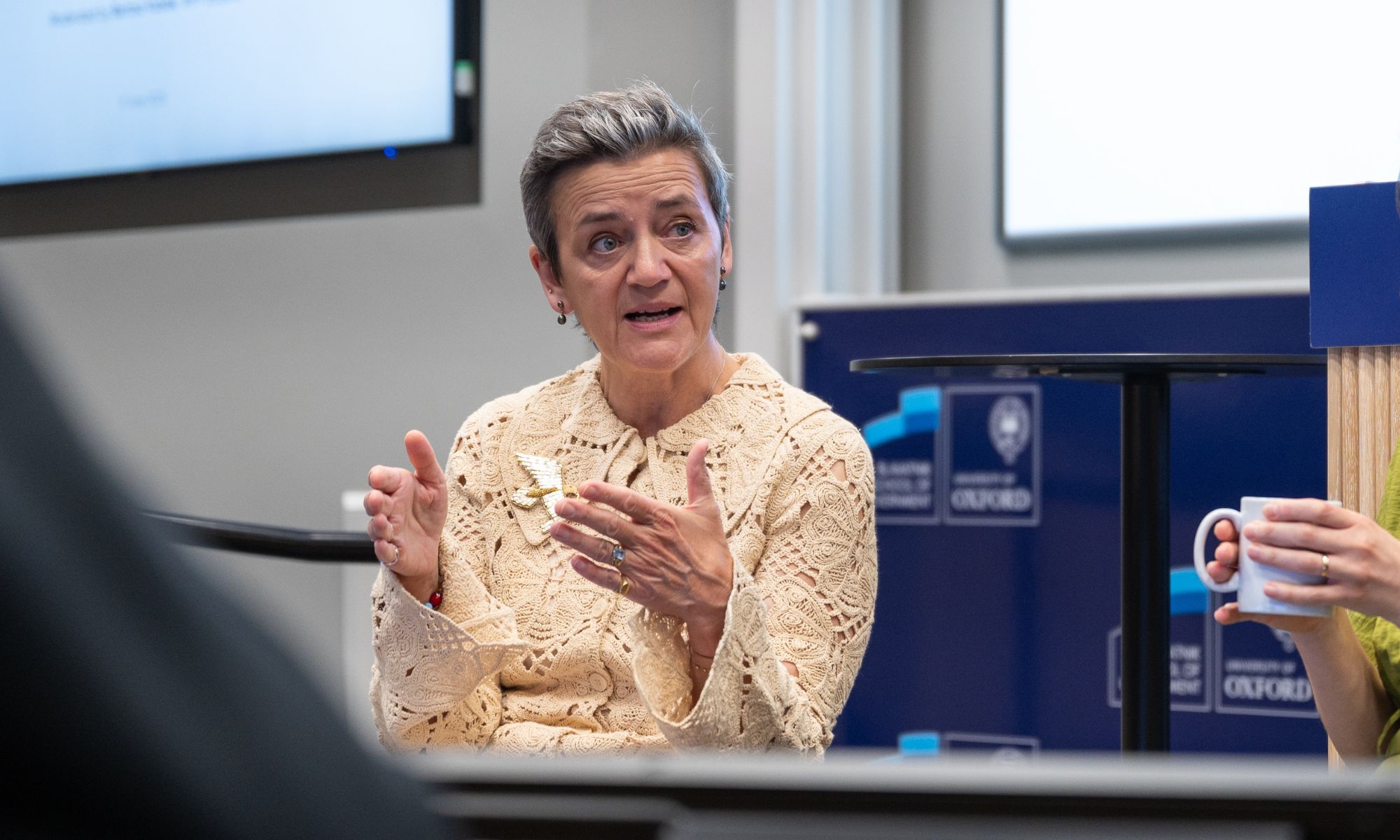 Margrethe Vestager speaking during a discussion at the Blavatnik School of Government, University of Oxford. She is gesturing with her hands while seated. A blurred audience co-panelist holding a cup held is visible in the foreground.
