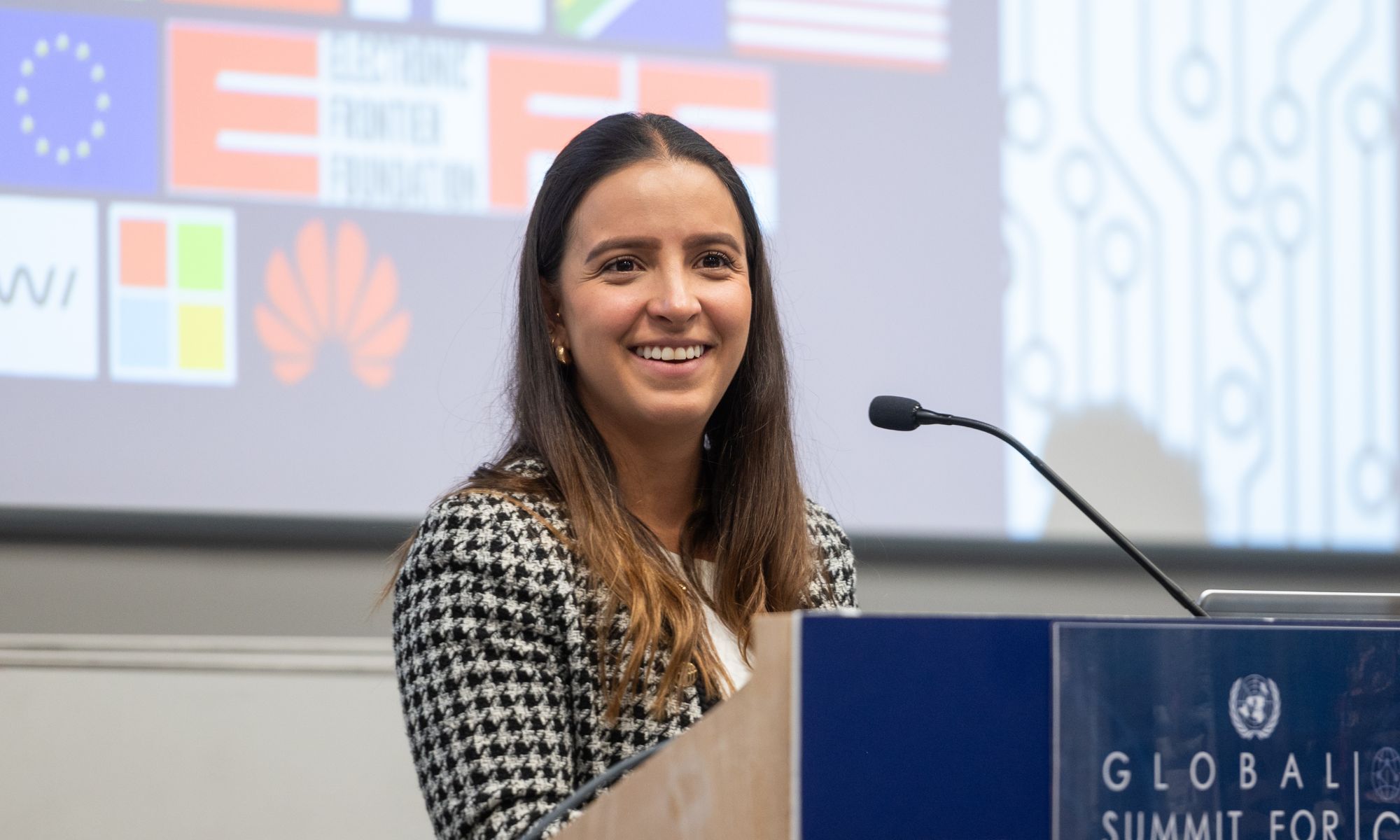 Female student is smiling at a lectern. Behind her some flags are displayed on a projector.