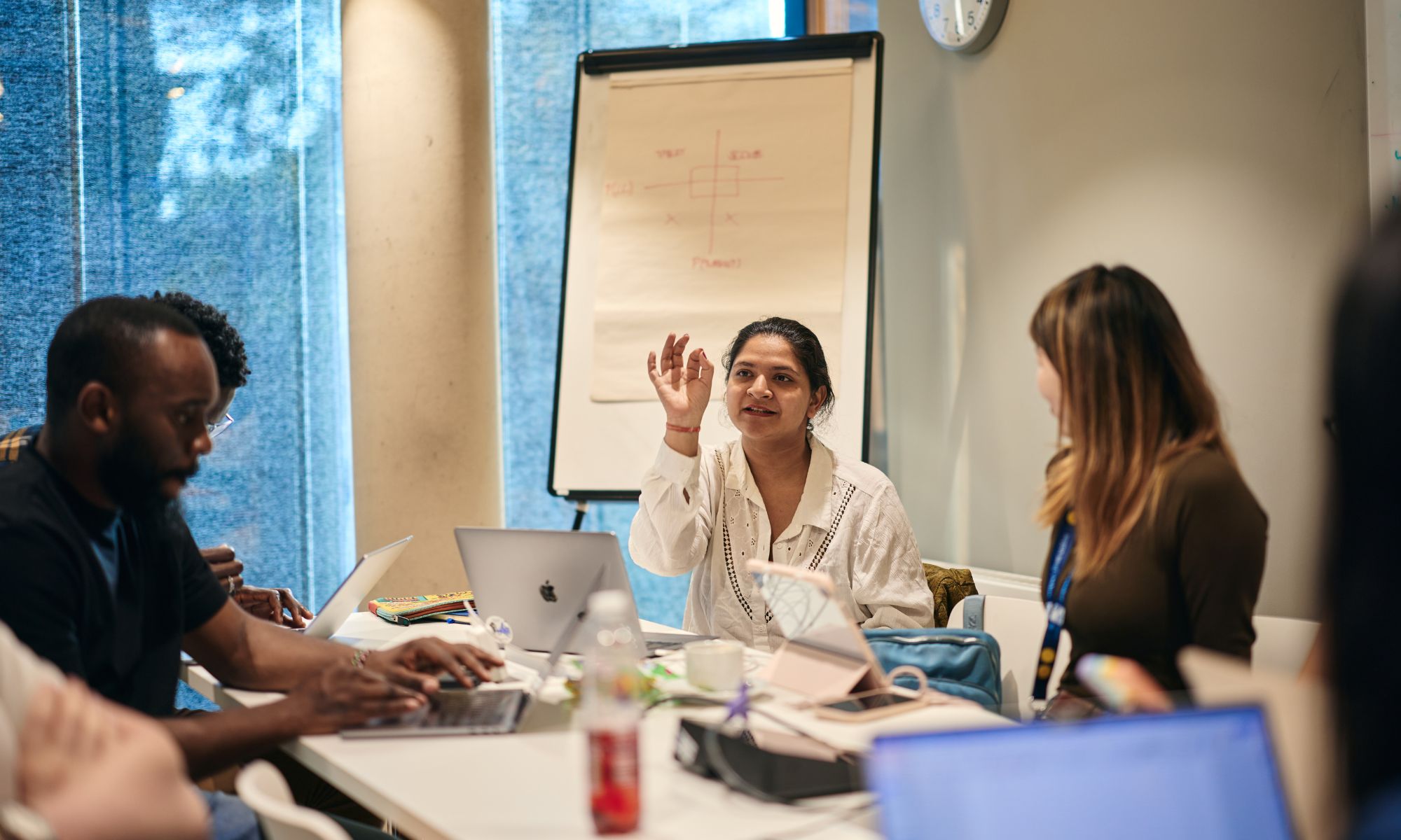 A female student sits at a table with other students in front of a flip chart. She has her hand in the air as though she is describing an idea.