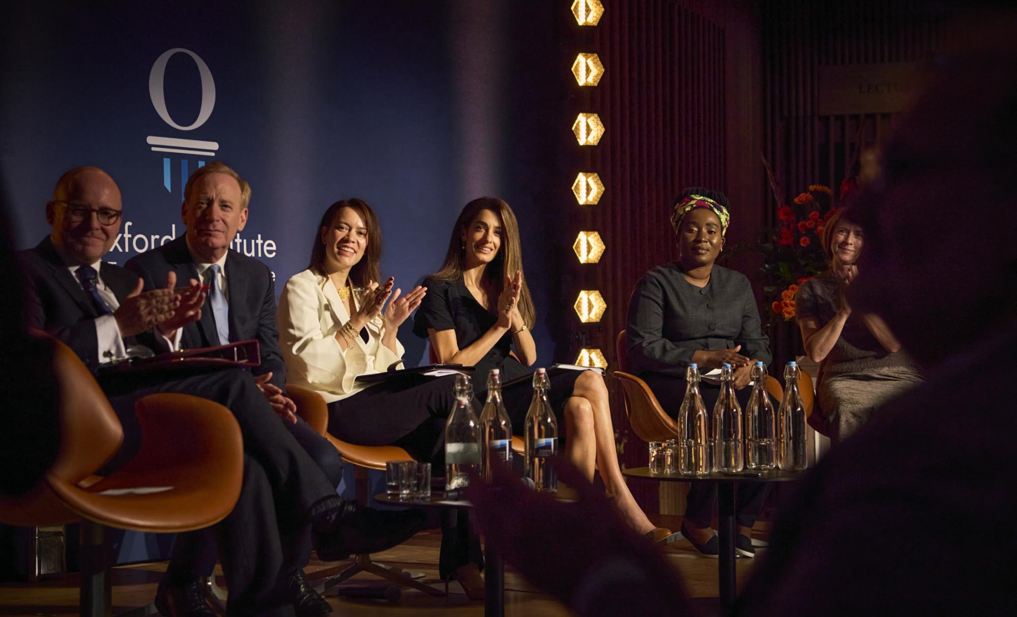Panel of Lord Herner, Microsoft CEO Brad Smith, Philippa Webb and Amal Clooney smiling and clapping against a branded backdrop