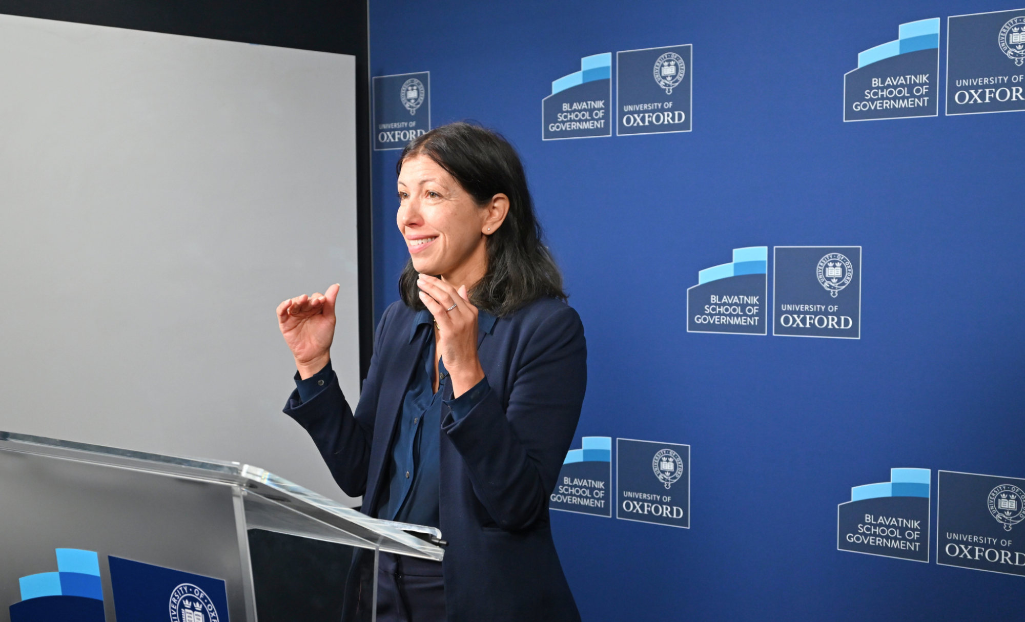 Professor Maya Tudor stands at a lecturn teaching in a green room with a branded Blavatnik School backdrop.