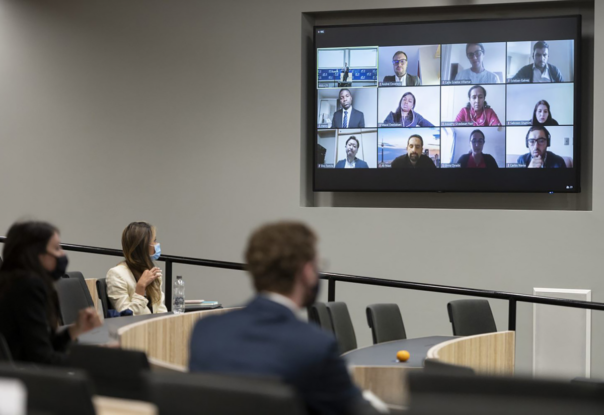 Three students sit socially distanced in a lecture theatre, facing the front where a screen displays other members of the class participating remotely