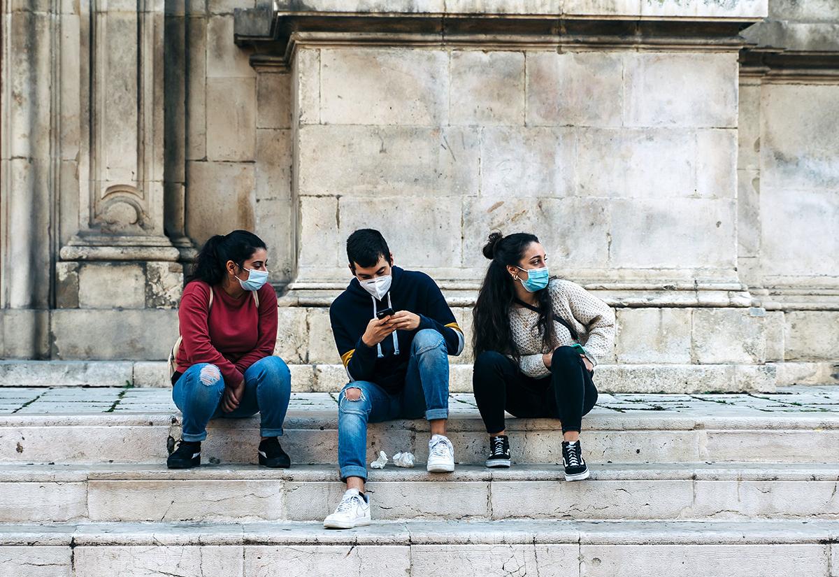 Three Italian teenagers with surgical masks chat on the marble steps of a church in Abruzzo, Italy