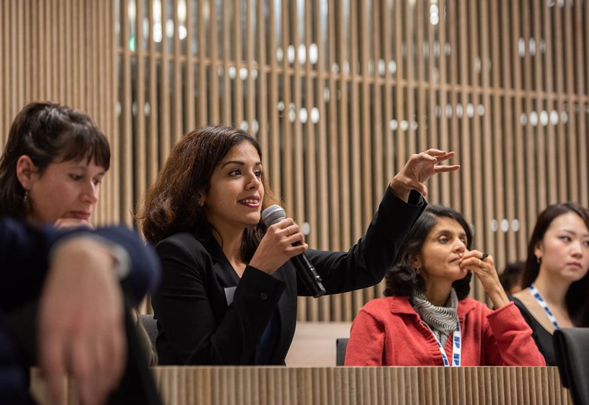 During a conference, a woman asks a question with a microphone in her right hand and raises her left hand