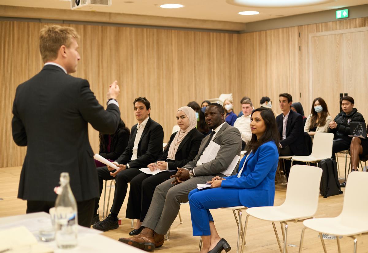 Faculty member Dr Tom Simpson talks to a group of 16 students sitting in rows in front of him