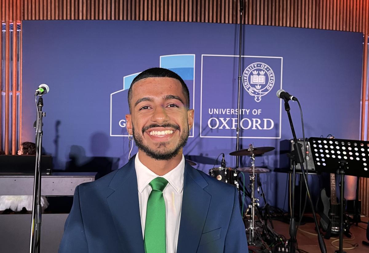 Photo of Giancarlo smiling in a blue suit and green tie against a background of a screen with the University of Oxford logo
