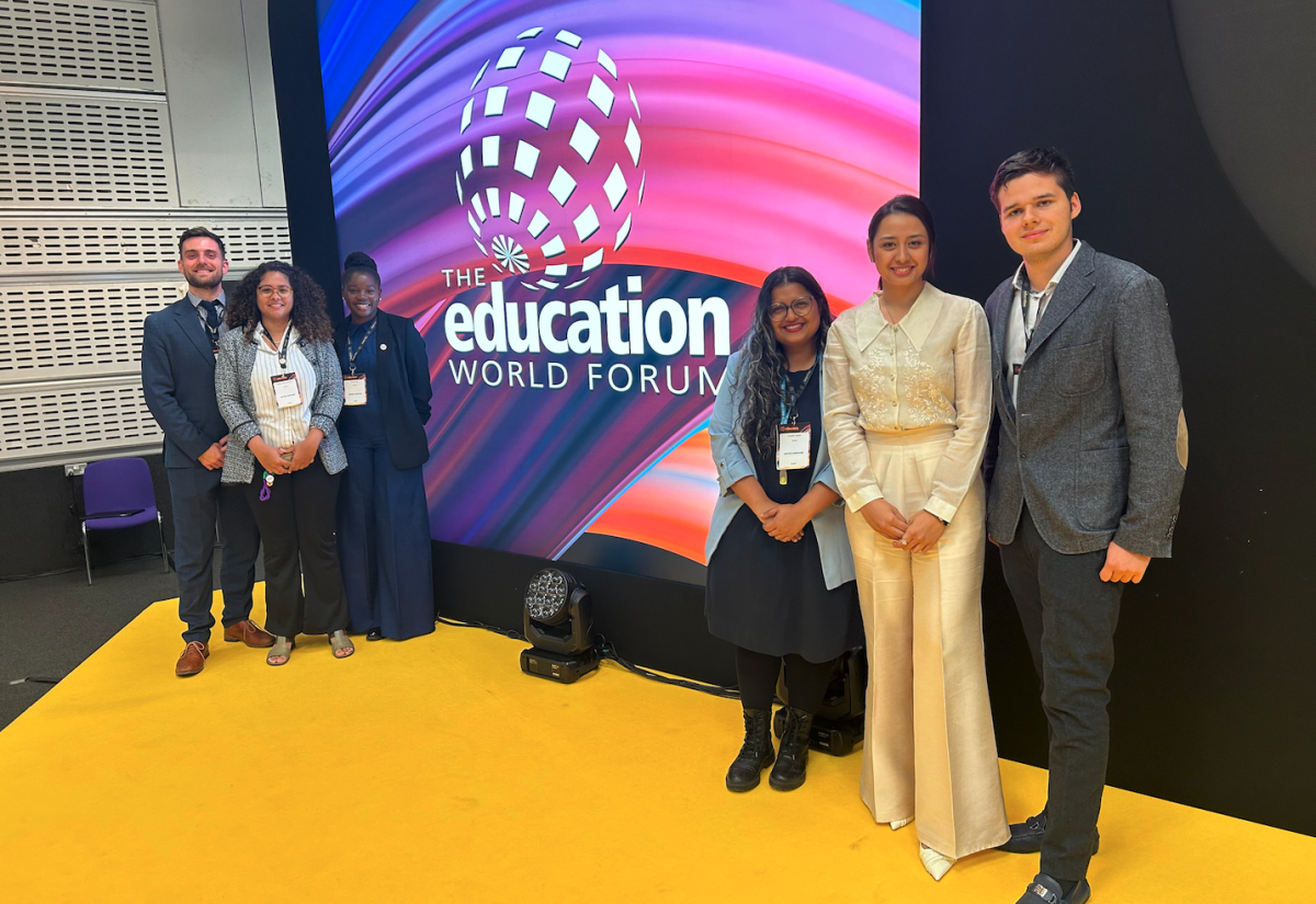 Photo of the Education World Forum logo on a screen with stripes purple and pink stripes and six smiling people in suits on either side of the logo