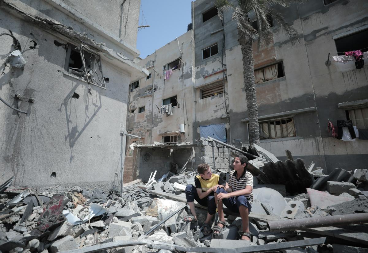 Two boys sit on rubble in Gaza, surround by buildings which have been bombed.