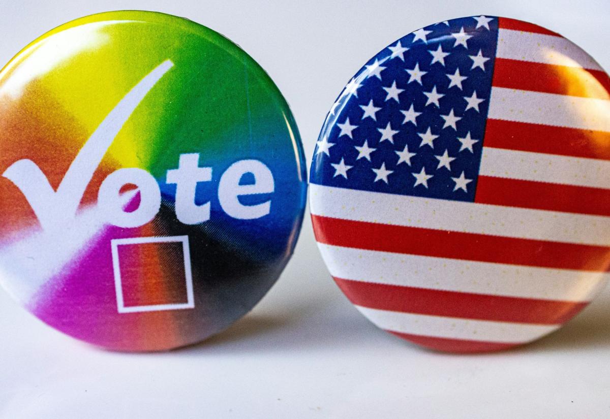 Two round badges side by side. The left badge has a rainbow background with the word "Vote" in white letters and a check box below it, symbolising LGBTQ+ pride and voting. The right badge features the design of the United States flag.