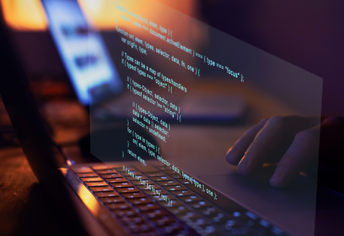 A close-up of a person typing on a laptop keyboard with lines of code projected or digitally superimposed above the keyboard. The background is softly blurred, showing another screen and a dimly lit workspace, creating a focused and tech-centric atmosphere.