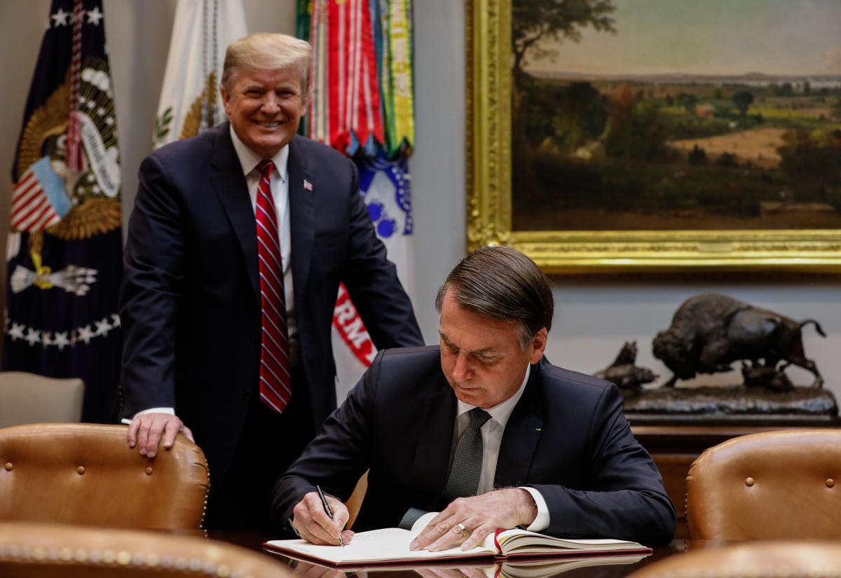 President of Brazil sits at a desk signing a document. President Trump is stood behind him smiling.