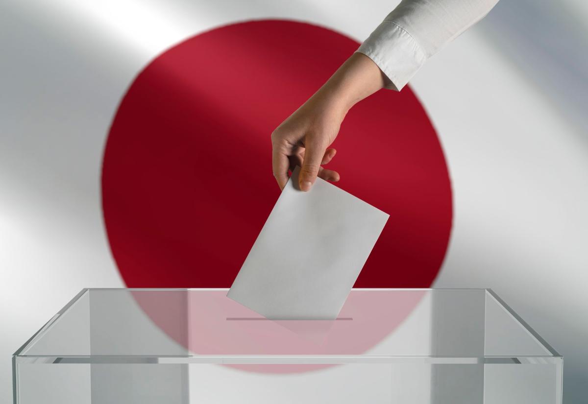 A hand is putting a white voter slip into a ballot box in front of the Japanese flag.