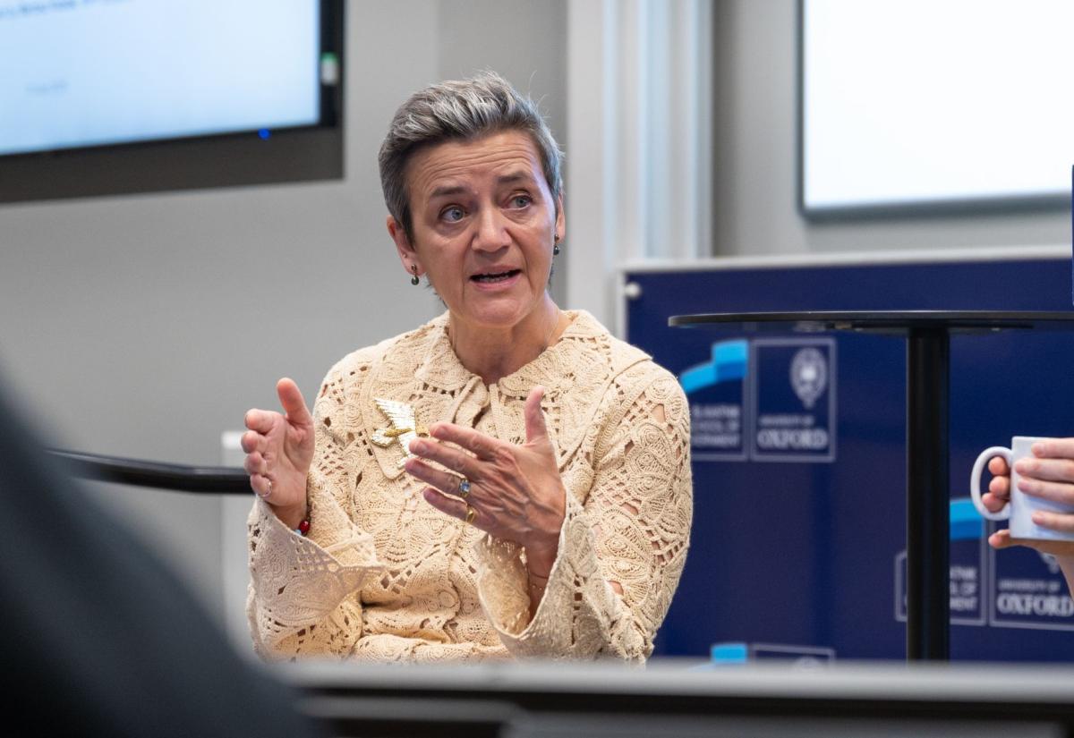 Margrethe Vestager speaking during a discussion at the Blavatnik School of Government, University of Oxford. She is gesturing with her hands while seated. A blurred audience co-panelist holding a cup held is visible in the foreground.