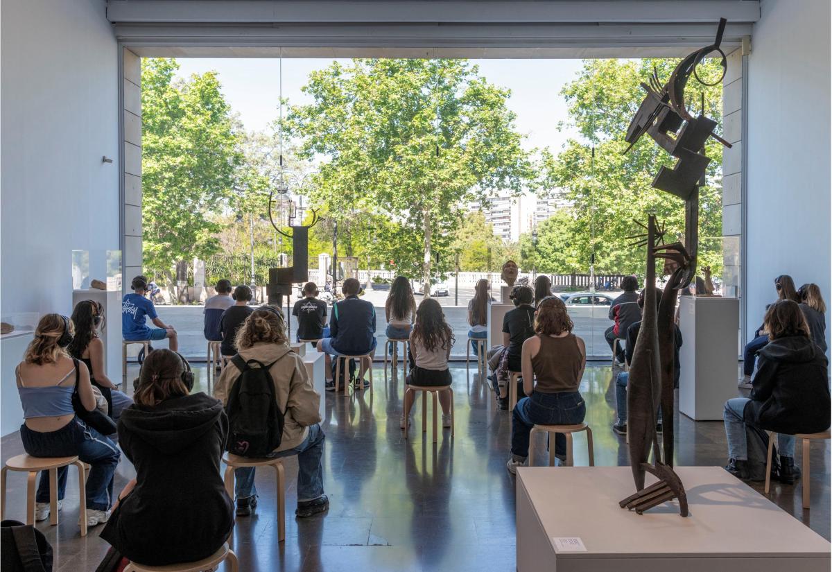 A group of people sat on chairs spaced out in a museum, looking out a window.