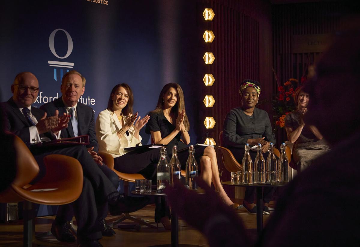 Panel of Lord Herner, Microsoft CEO Brad Smith, Philippa Webb and Amal Clooney smiling and clapping against a branded backdrop