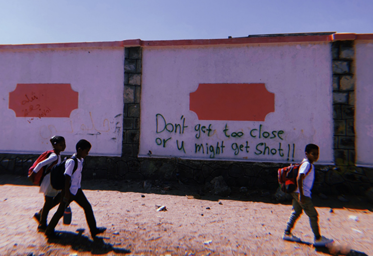Children walk past a graffitied wall in Aden, Yemen. The graffiti reads 'Don't get too close or u might get shot!!'.