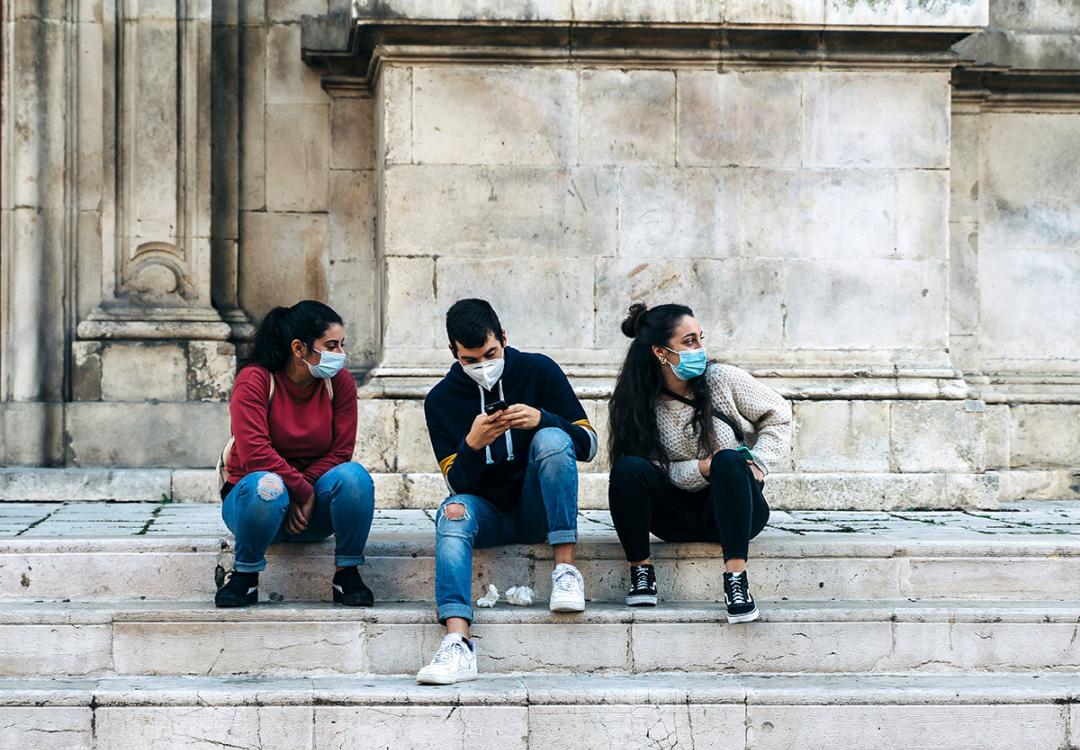 Three Italian teenagers with surgical masks chat on the marble steps of a church in Abruzzo, Italy
