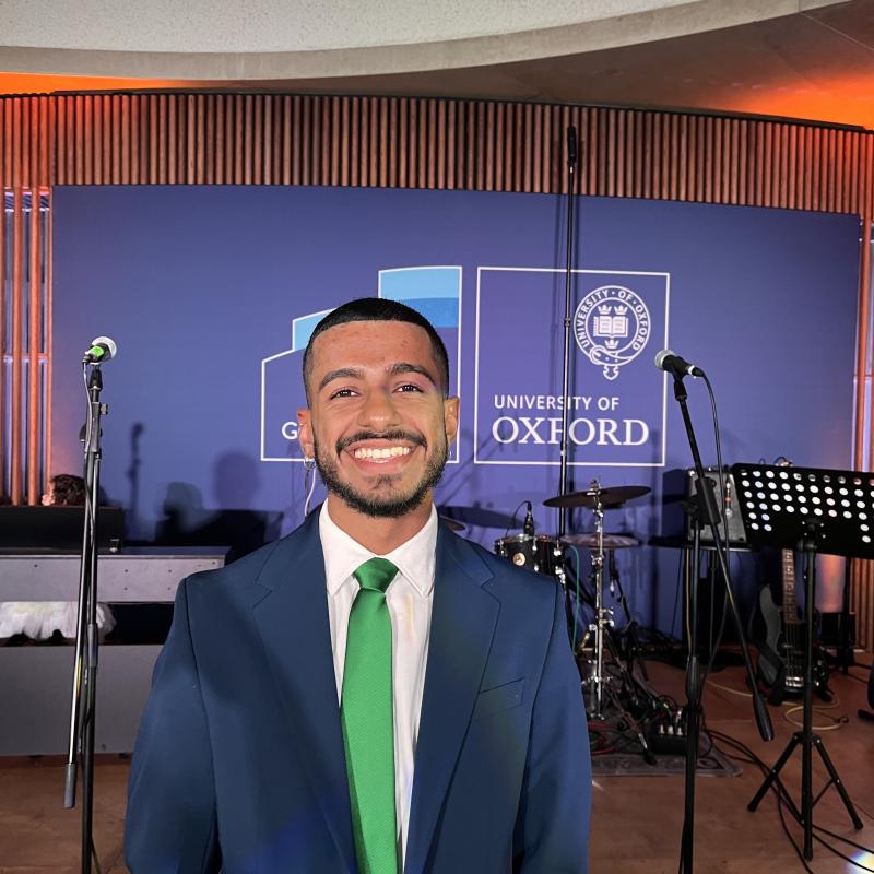 Photo of Giancarlo smiling in a blue suit and green tie against a background of a screen with the University of Oxford logo