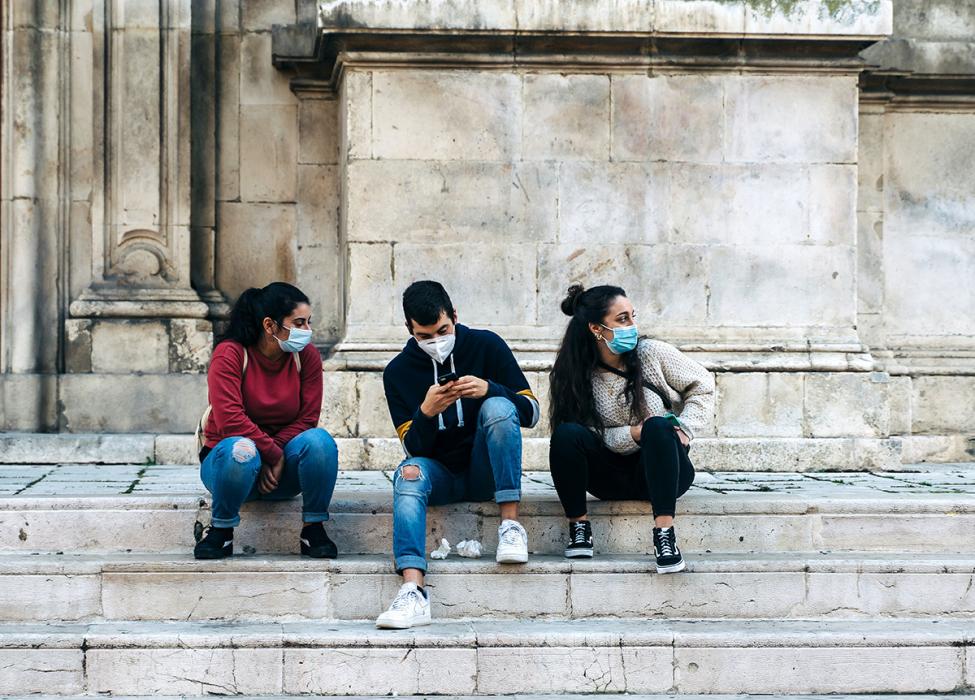 Three Italian teenagers with surgical masks chat on the marble steps of a church in Abruzzo, Italy