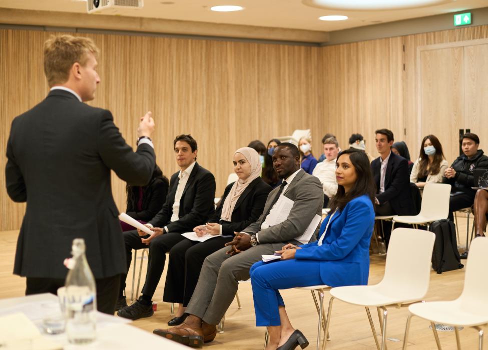 Faculty member Dr Tom Simpson talks to a group of 16 students sitting in rows in front of him